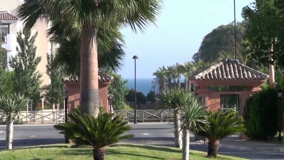 Frame from the complex tour video: palm-lined entrance roundabout with terracotta gatehouses and the sea in the distance